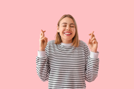 Happy young woman in striped sweatshirt crossing fingers on pink background