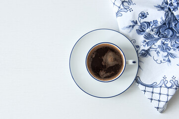 White cup of coffee with saucer, blue and white napkins on a white table. View from above.