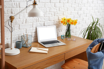 Workplace with modern laptop and beautiful narcissus flowers near white brick wall