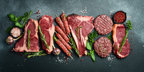 A set of juicy raw steaks, kebabs, cutlets and meat with spices and herbs. On a black stone background. Top view. - obrazy, fototapety, plakaty