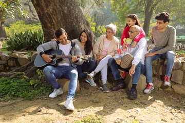 Happy Indian big family sitting on a pedestal under the shades of a tree and watching a family member playing guitar.