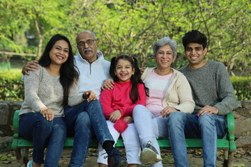Fototapeta premium Indian happy family having a good cheerful time sitting on a bench in the green park surrounded with a serene atmosphere and fresh air environment in day time.