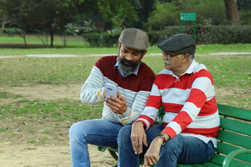 Father and mid aged son having a good time sitting on a park bench holding a mobile phone in hand. they are surrounded with lush greens and serene environment.