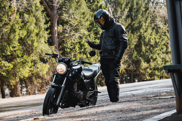 motorcyclist in a leather jacket and helmet on a custom motorcycle cafe racer on a forest road - obrazy, fototapety, plakaty
