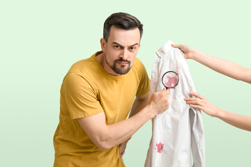 Handsome man with magnifier examining stained shirt on green background