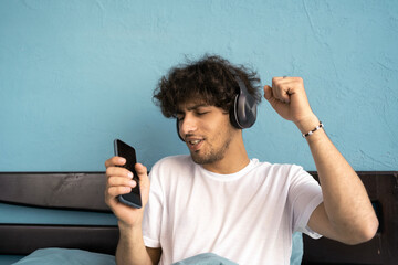 Young happy man wake up in morning listening music in headphones with smartphone, relaxing smiling, and dancing in bed