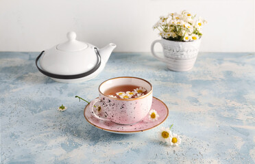 Teapot with cup of natural chamomile tea and flowers on blue grunge table near wall