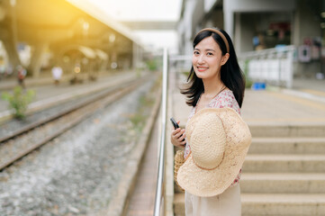 young asian woman traveler with weaving basket waiting for train in train station. Journey trip lifestyle, world travel explorer or Asia summer tourism concept.