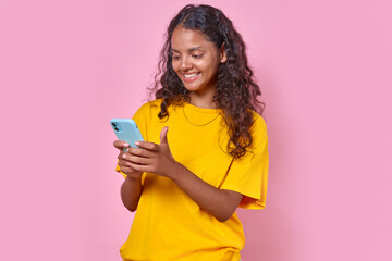 Young optimistic Indian woman with smartphone chatting via SMS or internet messenger for messaging and funny photos dressed in casual clothes stands in pink studio. Correspondence, communication