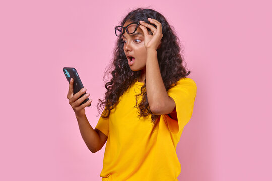 Young surprised Indian woman with phone raises glasses from eyes after seeing unexpected message from ex-boyfriend or reading news in yellow press stands on isolated pink background.
