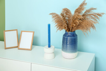 White chest of drawers with pampas grass, candle and blank pictures near blue wall in room, closeup