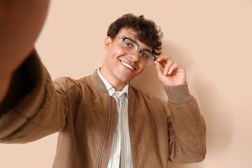 Young man in stylish eyeglasses taking selfie on beige background