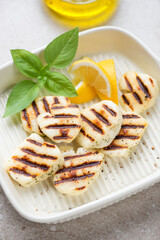 Slices of grilled haloumi cheese with lemon wedges and green basil on a beige serving tray, vertical shot, selective focus