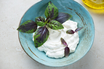 Ricotta cheese and fresh basil leaves in a turquoise bowl, horizontal shot on a light-beige stone...
