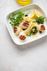 Serving tray with grilled scallops, spaghetti, lemon and fresh parsley, vertical shot on a light-beige stone background with space, elevated view