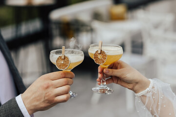Hands of a happy loving couple with cocktail glasses, celebrating wedding day at restaurant. Selective focus on drinks in hands of young couple