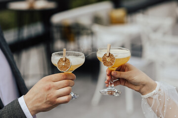 Close-up of hands of woman and man are clinking, cheers with glasses of cocktail. Couple celebrating wedding, anniversary with cocktails with orange. Refreshing alcoholic drink. - obrazy, fototapety, plakaty