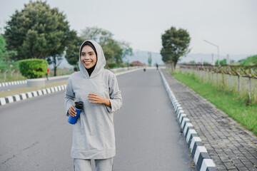 Fototapeta premium asian muslim woman running and exercising while holding drink bottle outdoors excitedly