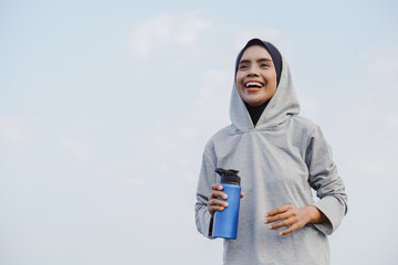 asian muslim woman running and exercising while holding drink bottle outdoors excitedly