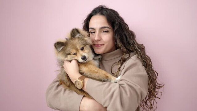 Young hispanic woman with dog smiling confident standing over isolated pink background