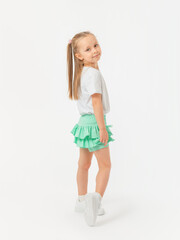 A 5-year-old girl with long hair is standing sideways in a T-shirt and holding on to her skirt, looking at the camera in sneakers on a white background.