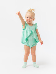 A blonde 2-year-old girl in a green dress and socks stands and points with her index finger at something with an expressive look on a white background.