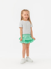 A cute 5-year-old girl in a T-shirt, sneakers and a green skirt with long hair smiles and looks at the camera on a white background. photo
