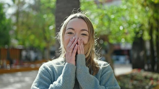 Young blonde woman standing with surprise expression covering mouth with hands at park