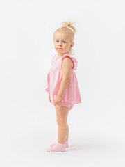 Excitement, sadness. A blonde girl 1-2 years old in a pink dress and socks stands sideways and looks at the camera on a white background.