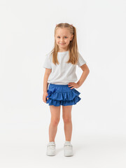 One hand on the waist. A girl smiling with long hair in a blue skirt, white T-shirt and sneakers stands on a white background expressively looking at the camera.