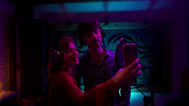 Positive woman holding smartphone and recording tik tok video while singing with man at neon light studio