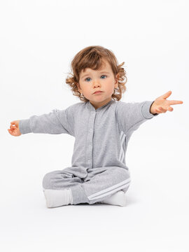 Surprise, question, disappointment. A 2-year-old toddler with curly hair in a gray jumpsuit and socks is sitting on the floor, arms outstretched and legs crossed on a white background.