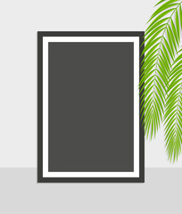 Empty black photo frame with palm leaves and shadow in front of white wall background.