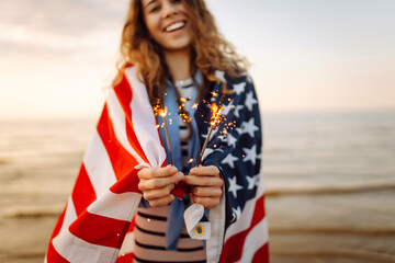 Young woman with American flag  and sparklers on the beach. Patriotic holiday. USA celebrate 4th of...