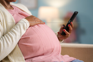 Close up shot of indian pregnant woman busy using mobile phonw by holding tummy at home - concept of digital connection, technology and Digital maternity