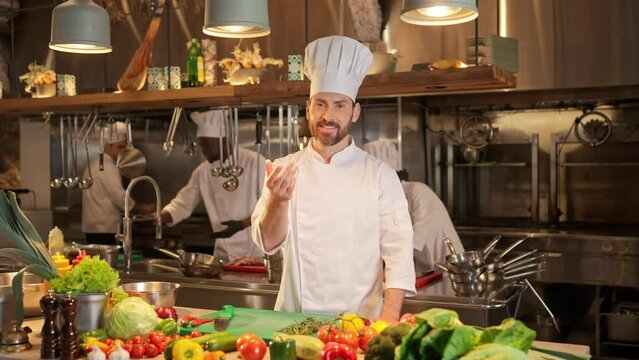 Portrait of bearded chef smiling and making traditional bon appetit gesture. Cook dressed in special uniform and white hat stands at background of restaurant professional kitchen.