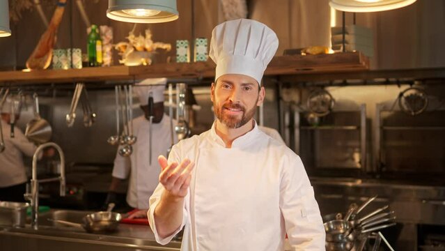 Portrait of bearded man cook in special white uniform and hat giving chef's kiss at restaurant professional kitchen. Male chef stands near work place, looking at camera and smiling.