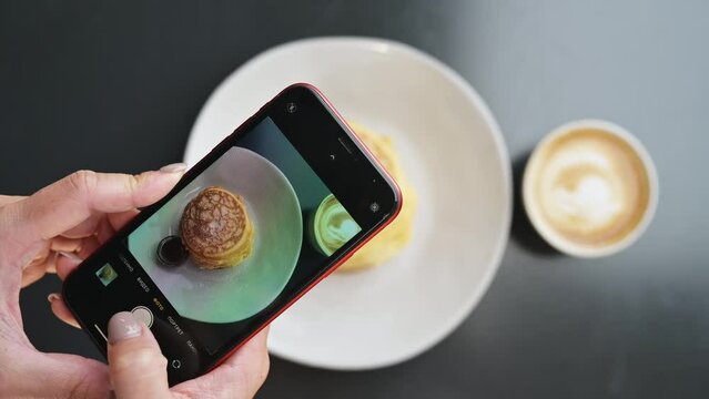 Top view of female hands holding a smartphone, a woman influencer blogger, taking pictures of food with her mobile phone for social media, taking pictures of pancakes and coffee before eating them