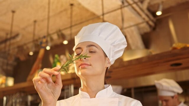Close-up portrait of female chef standing in restaurant professional kitchen and inhales smell of fragrant herbal spices. Woman dressed in special uniformvselects ingredients for preparation of dish.