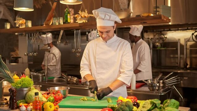 Portrait of cook in uniform shredding green fresh cucumber for prepearing salad or stew with sharp kitchen knife. Chef cooking delicious food at professional restaurant kitchen.