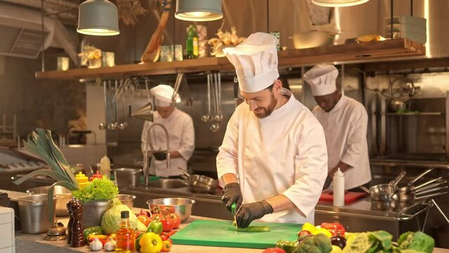 Smiling cook in uniform shredding green fresh cucumber for prepearing salad with sharp kitchen knife. Joyful chef cooking delicious food at professional restaurant kitchen.