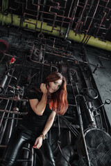 Fototapeta na wymiar Pensive young cover lady model in black posing in dark industrial room, thought looking at camera. Lovely redhead woman actress in industry premises. Fashionable stylish concept. Copy ad text space
