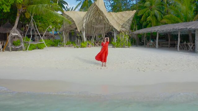 Beautiful sexy girl in a red dress walking down the white sand beach on a paradise tropical island on bamboo bungalows background on Thinadhoo, Maldives. Aerial drone view 4K.