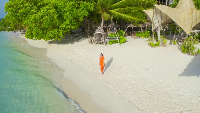 Beautiful sexy girl in a red dress walking down the white sand beach on a paradise tropical island on bamboo bungalows background on Thinadhoo, Maldives. Aerial drone view 4K.