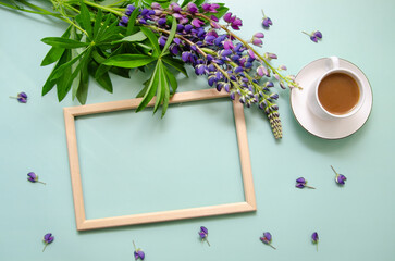 White cup of tea with lupine flowers and petals with empty wooden frame on blue background. Space for text or design.