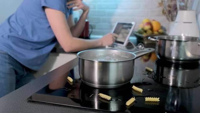 Young caucasian woman searching for video cooking recipe in tablet. Boiling water in saucepan on black induction cooking. Modern kitchen, household appliances, cookware. Girl use gadget, search recipe