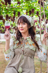 Woman wearing cute dress sitting on a swing holding glass of wine in vineyard smiling happily