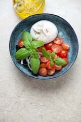 Blue bowl with burrata, red cherries, black olives and fresh basil, elevated view on a beige stone background, vertical shot with space