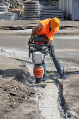A builder in an orange cape is ramming a trench with a vibratory rammer at a construction site.