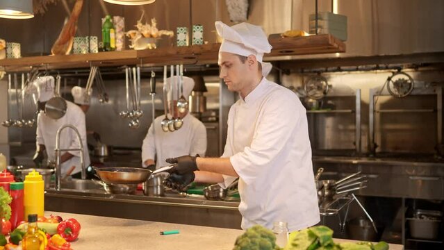 Portrait of professional master chef frying pieces of meat in pan. Adult cook dressed in special uniform preparing delicious dishes by prescription in restaurant commertial kitchen.